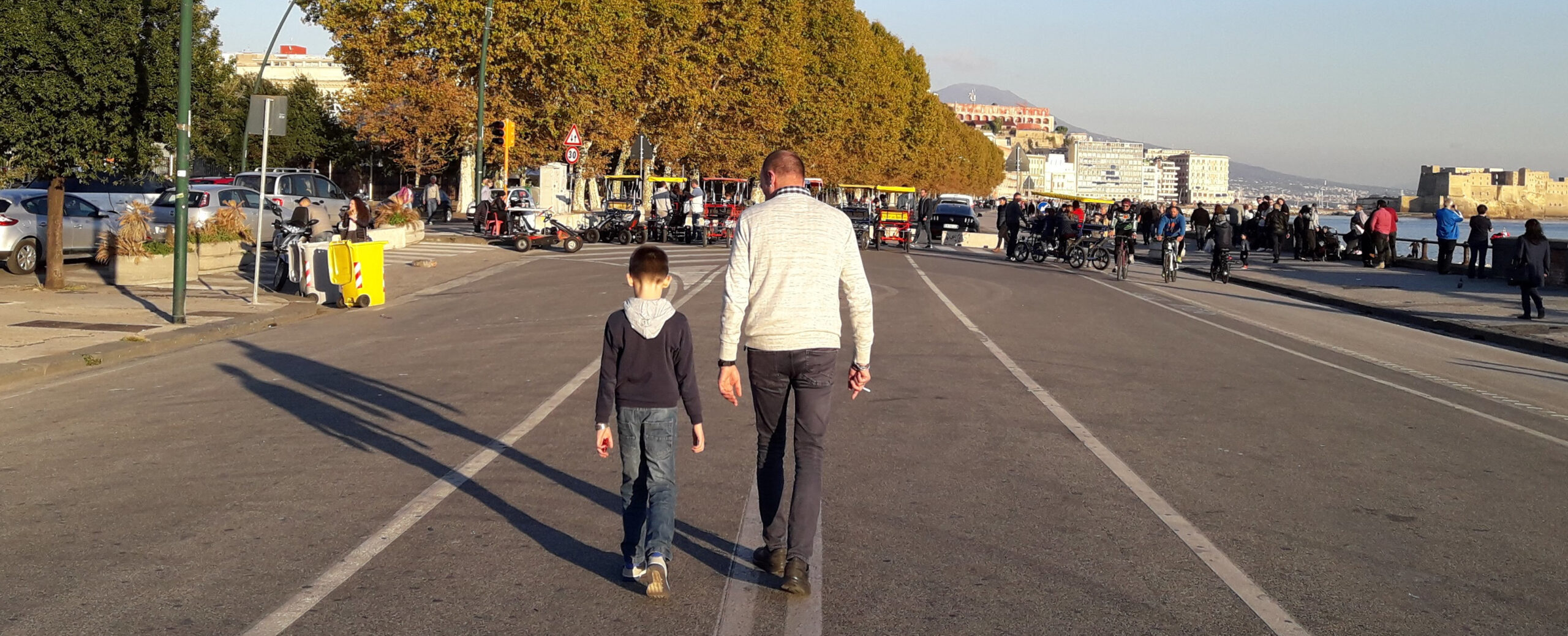 Un adulto e un bambino camminano di spalle sul lungomare, nella luce del pomeriggio.